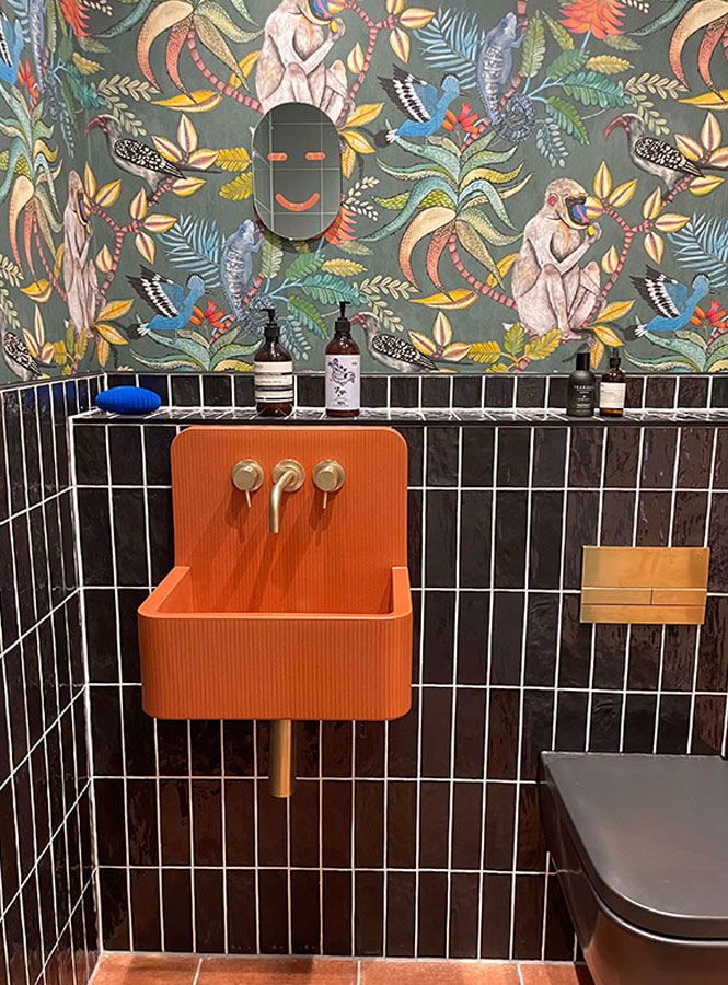 Orange concrete sink with gold taps, sits on vertical black gloss tiles with vibrate tropical and animal cole and son wallpaper