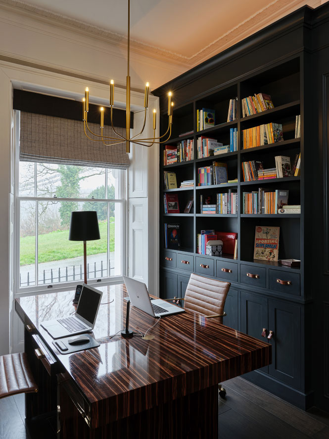 Desk with lamp on sits infront of a large window with dark painted bookshelves behind. There is a simple gold chandelier above the desk.