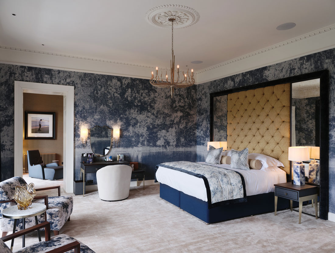 Master bedroom with large scale blue and white wallpanels. Large gold headboard with mirrors either side and bedside tables with marbled blue lamps